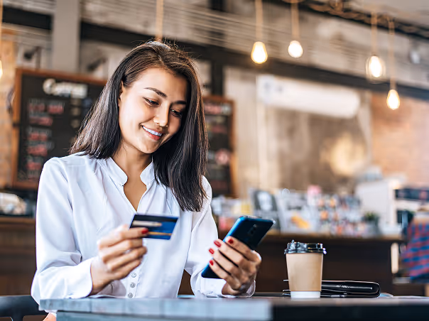 Smiling woman holding a credit card and smartphone, sitting at a table with a takeaway coffee cup in a cafe.