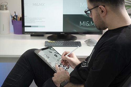 A graphic designer with glasses sits at a white desk, drawing logo sketches on a tablet