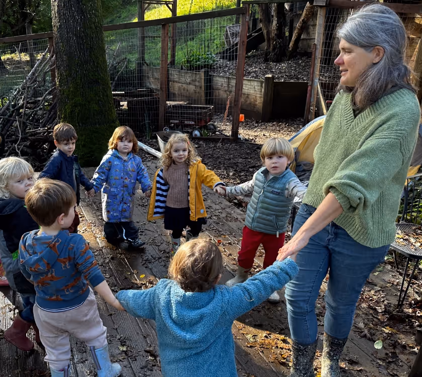 Teacher Teresa leading a circle game outdoors with a group of preschool children holding hands.