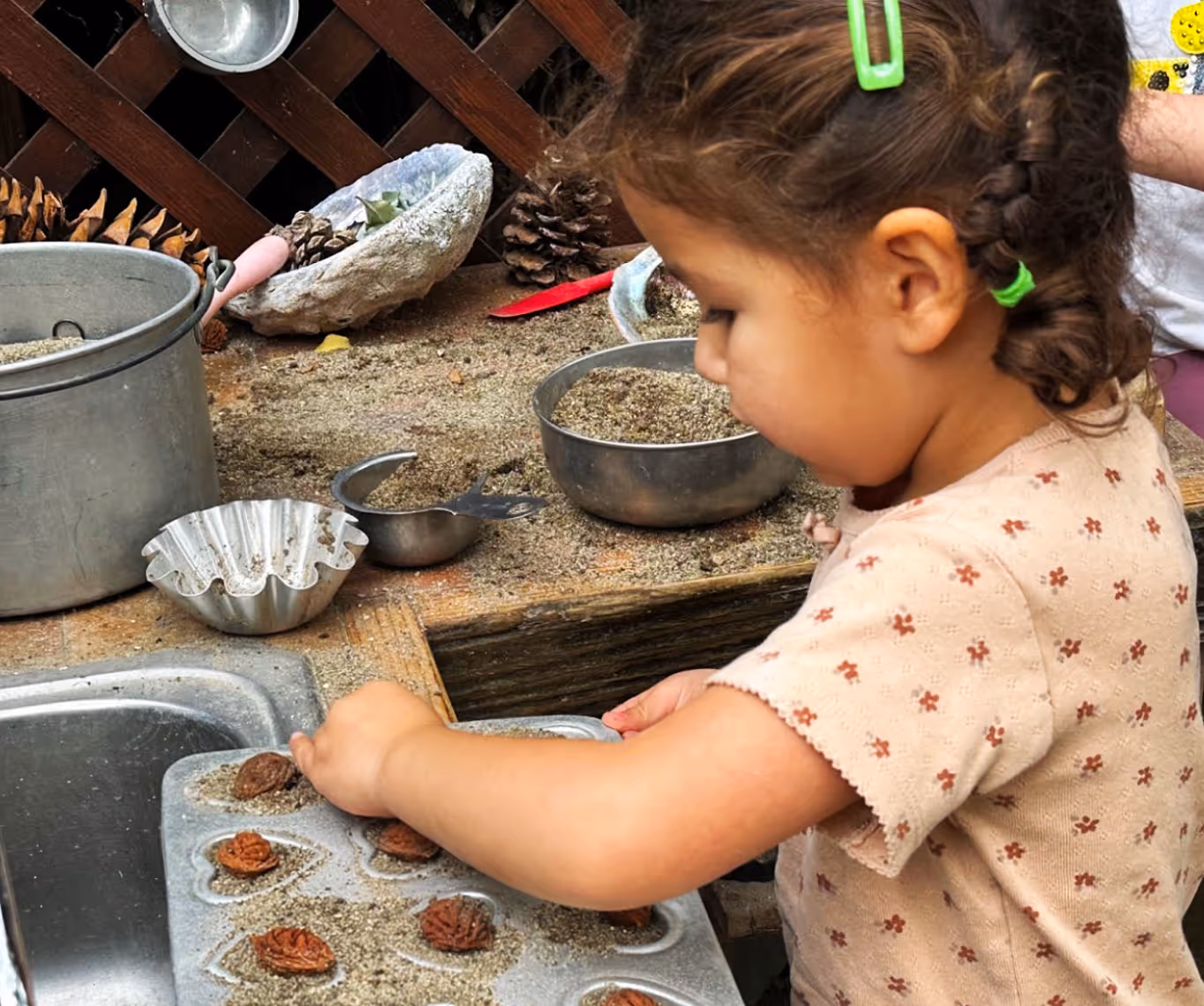 Young child playing with sand and natural objects in metal baking molds at a wooden mud kitchen.