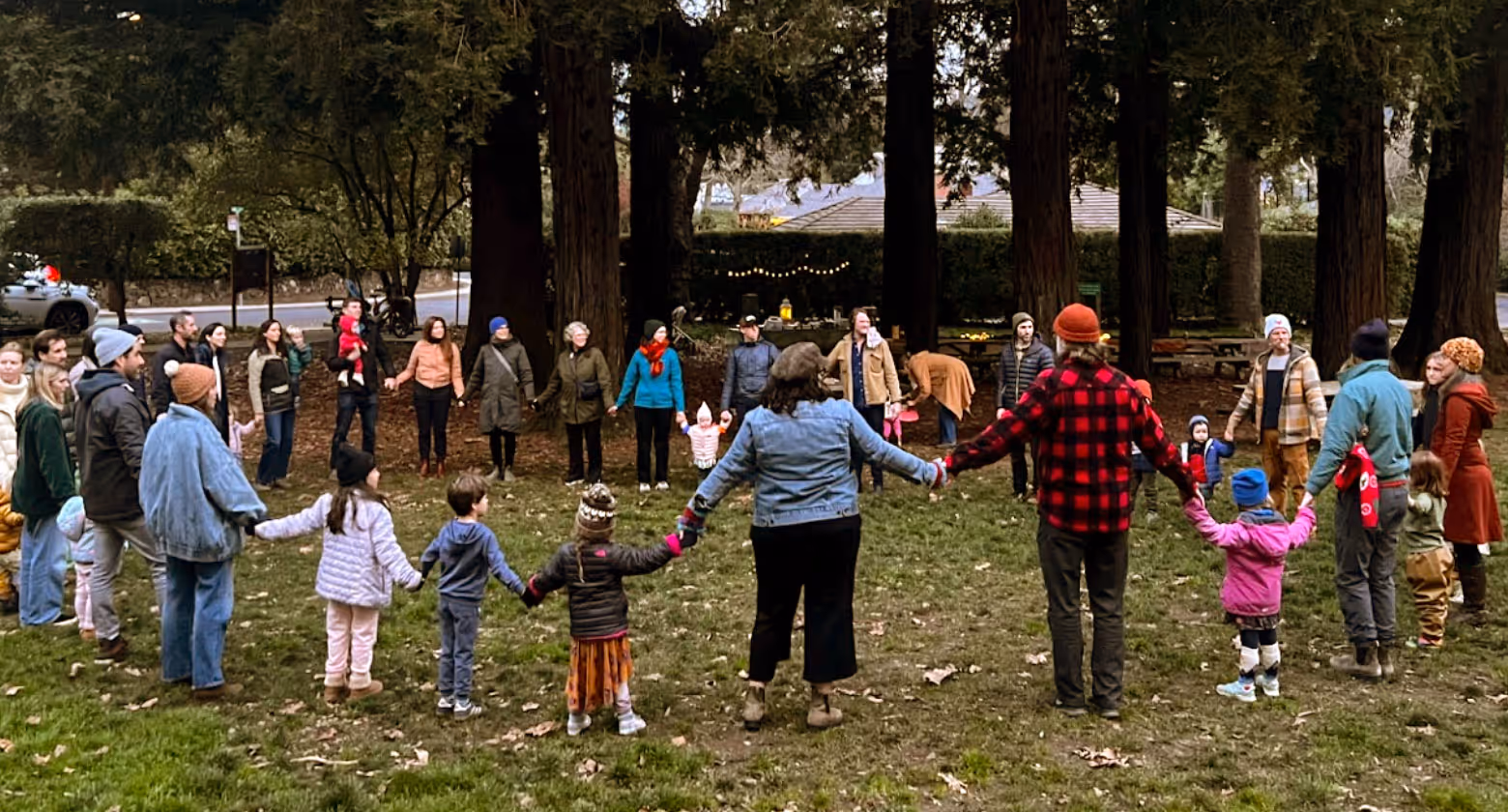 Group of adults and children standing in a circle holding hands in a park with tall trees.