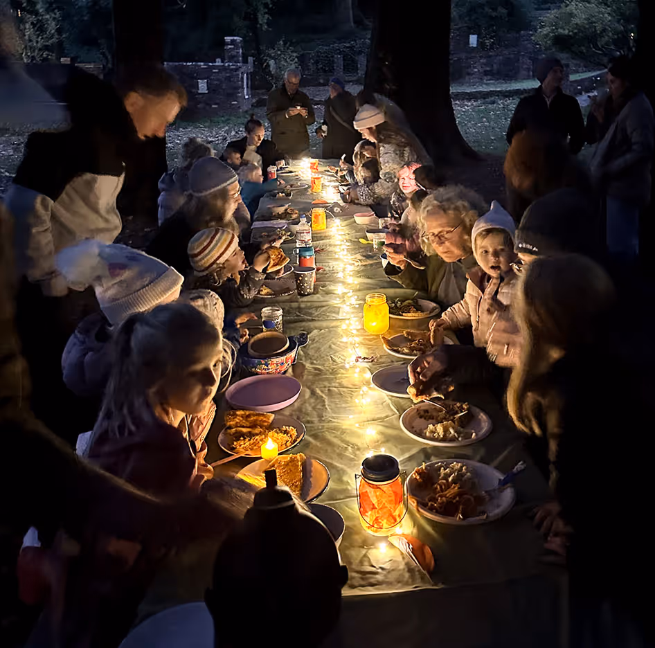 Large group of people, including children and adults, gathered around a long outdoor table with string lights and lanterns, sharing a meal at night.