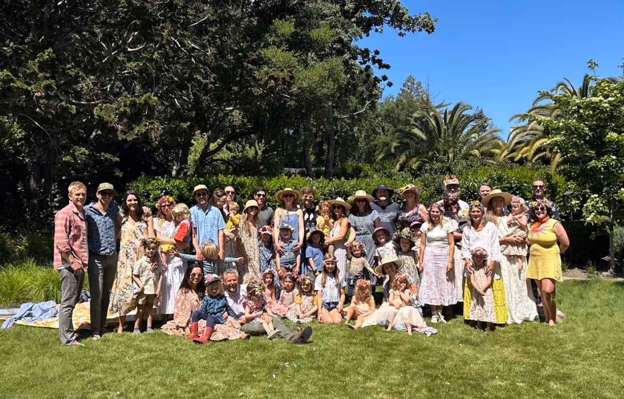 Large group of adults and children posing outdoors on grass, many wearing flower crowns and summer clothes under a clear blue sky.