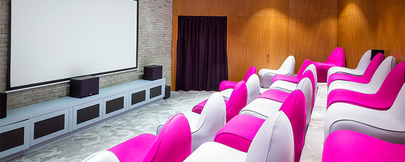 Modern conference room with white and pink cushioned seats facing a large projector screen on a brick wall.