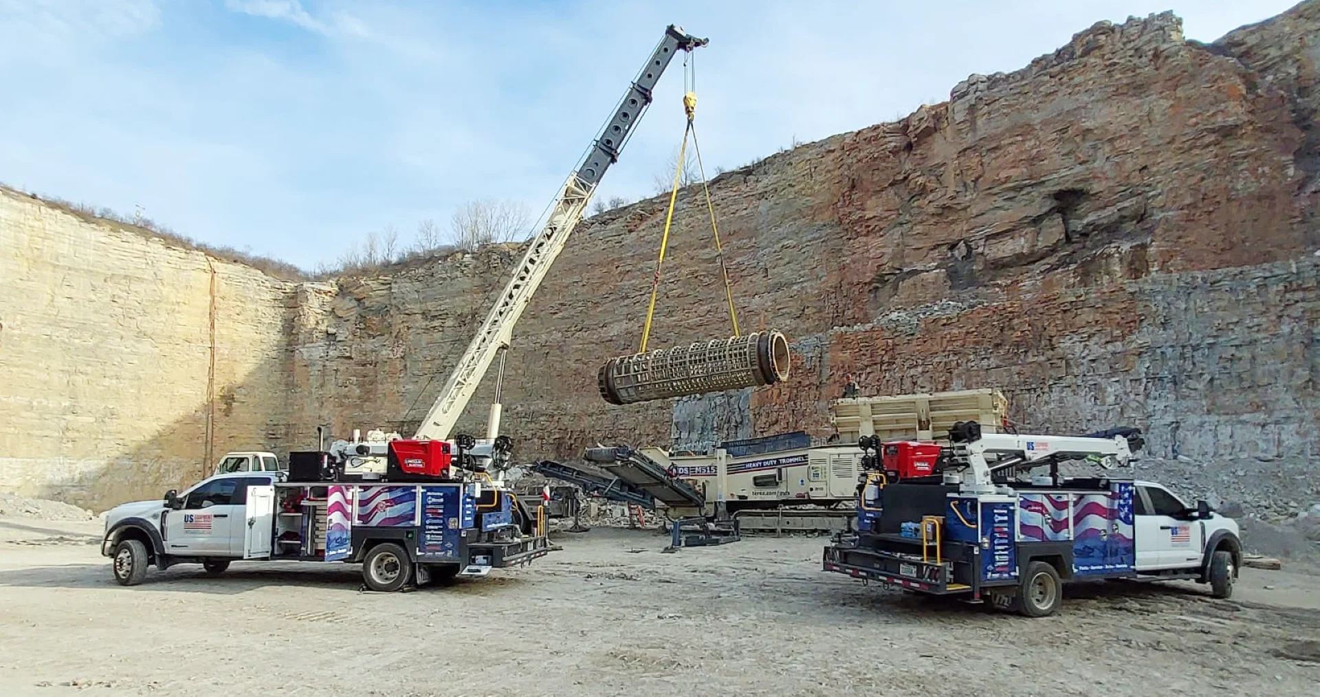 US Equipment Sales service trucks and crane lifting a heavy trommel drum inside a rock quarry, showcasing field service support for crushing and screening equipment.