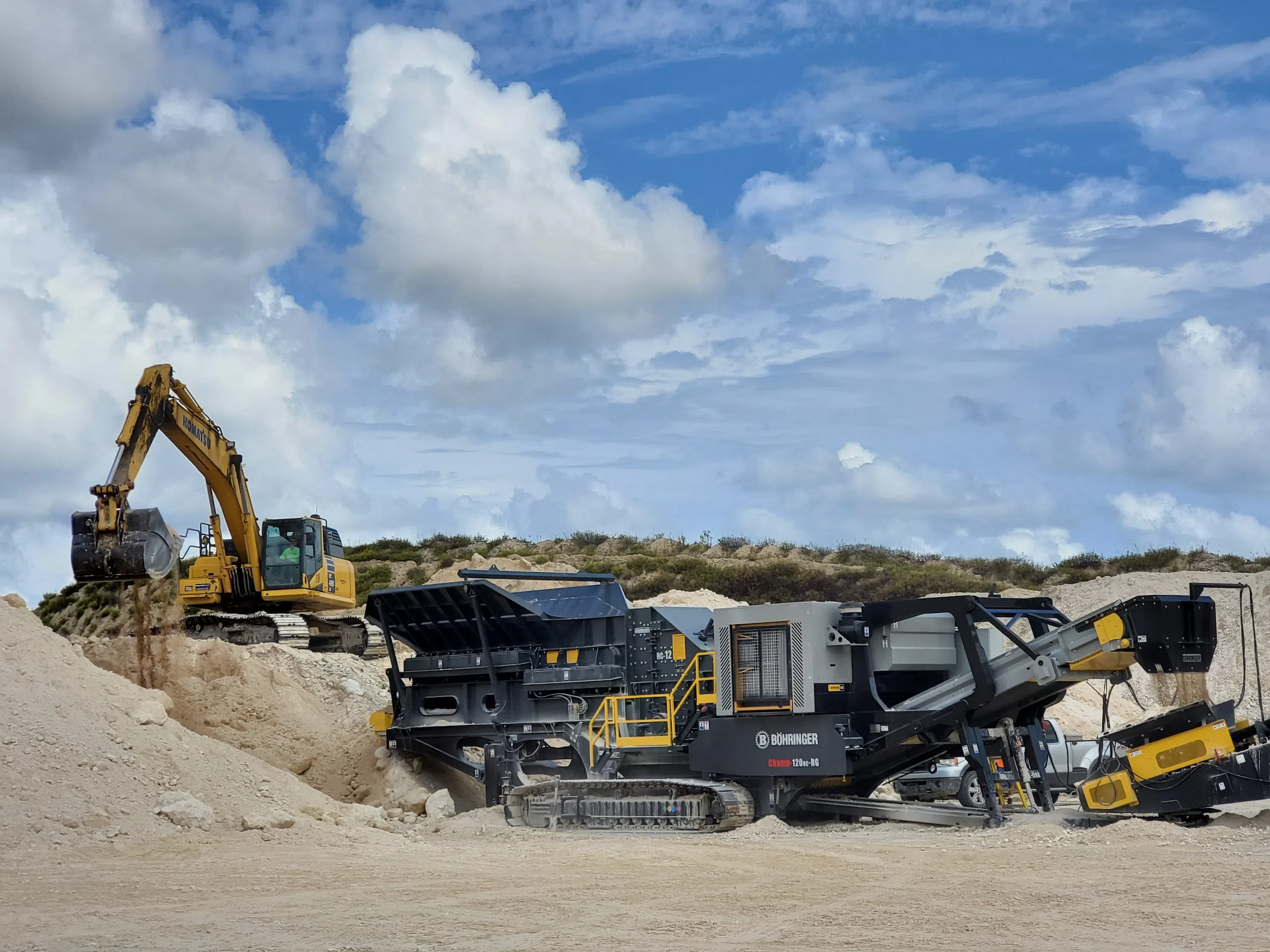 Böhringer mobile impact crusher processing material as a Komatsu excavator loads sand and rock into the feed hopper at a quarry site.