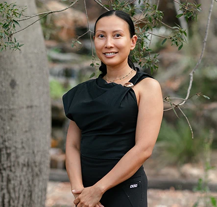 Woman in a black sleeveless top and leggings standing outdoors under leafy branches, hands clasped, smiling at the camera.