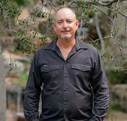 Man in a dark button-up shirt and jeans standing outdoors beneath leafy branches, hands resting on his thighs, looking at the camera.