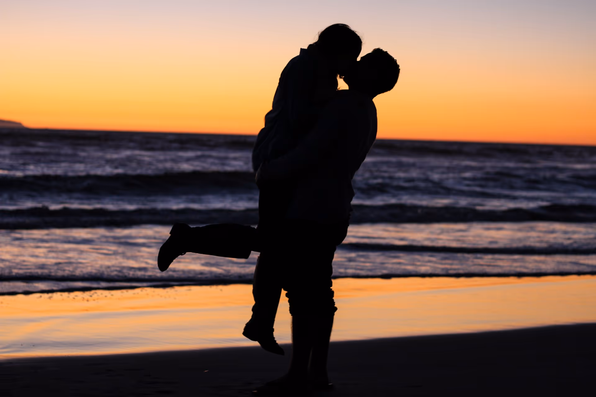 Silhouette of a couple embracing and kissing on a beach at sunset, with waves in the background and a colorful sky creating a romantic atmosphere.