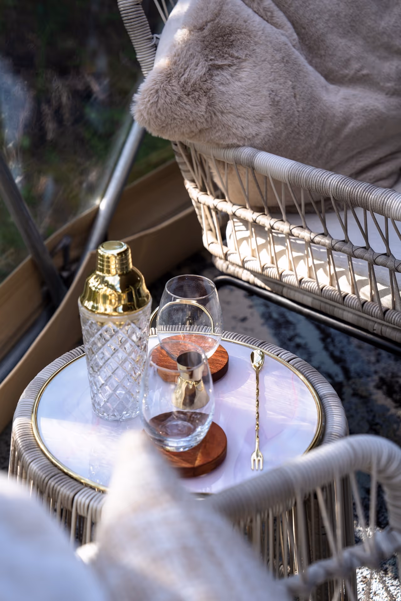 Cozy outdoor setting with wicker chairs, plush beige cushions, and a small round table. A glass cocktail shaker and glasses rest beside a golden spoon.