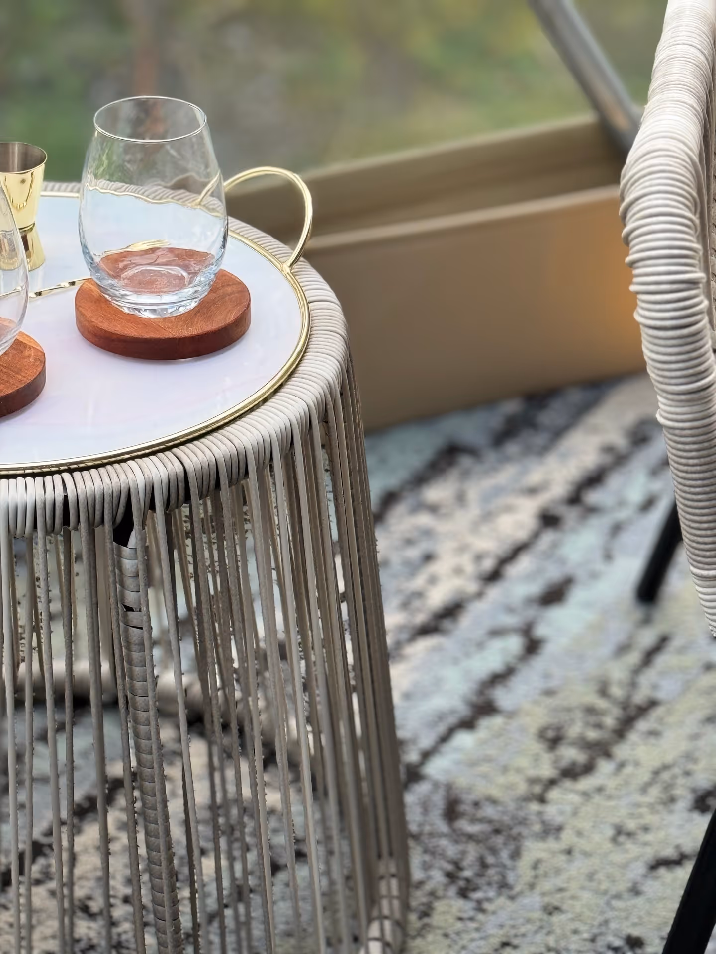 A round, woven side table holds two glasses on wooden coasters, adjacent to a patterned rug and chair. The scene is cozy and inviting near a window.