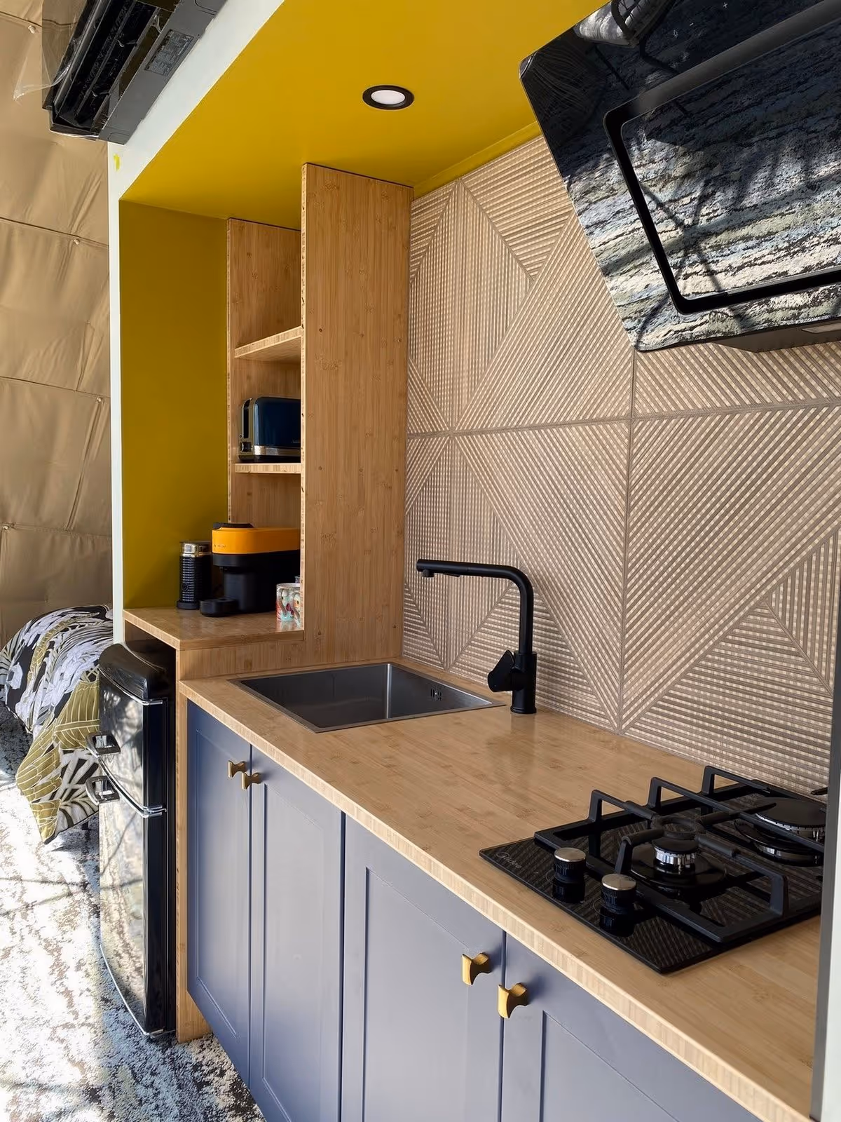 Modern kitchen with yellow walls, geometric backsplash, and wooden accents. Features include a gas stove, black faucet, and mini fridge. Cozy and chic.