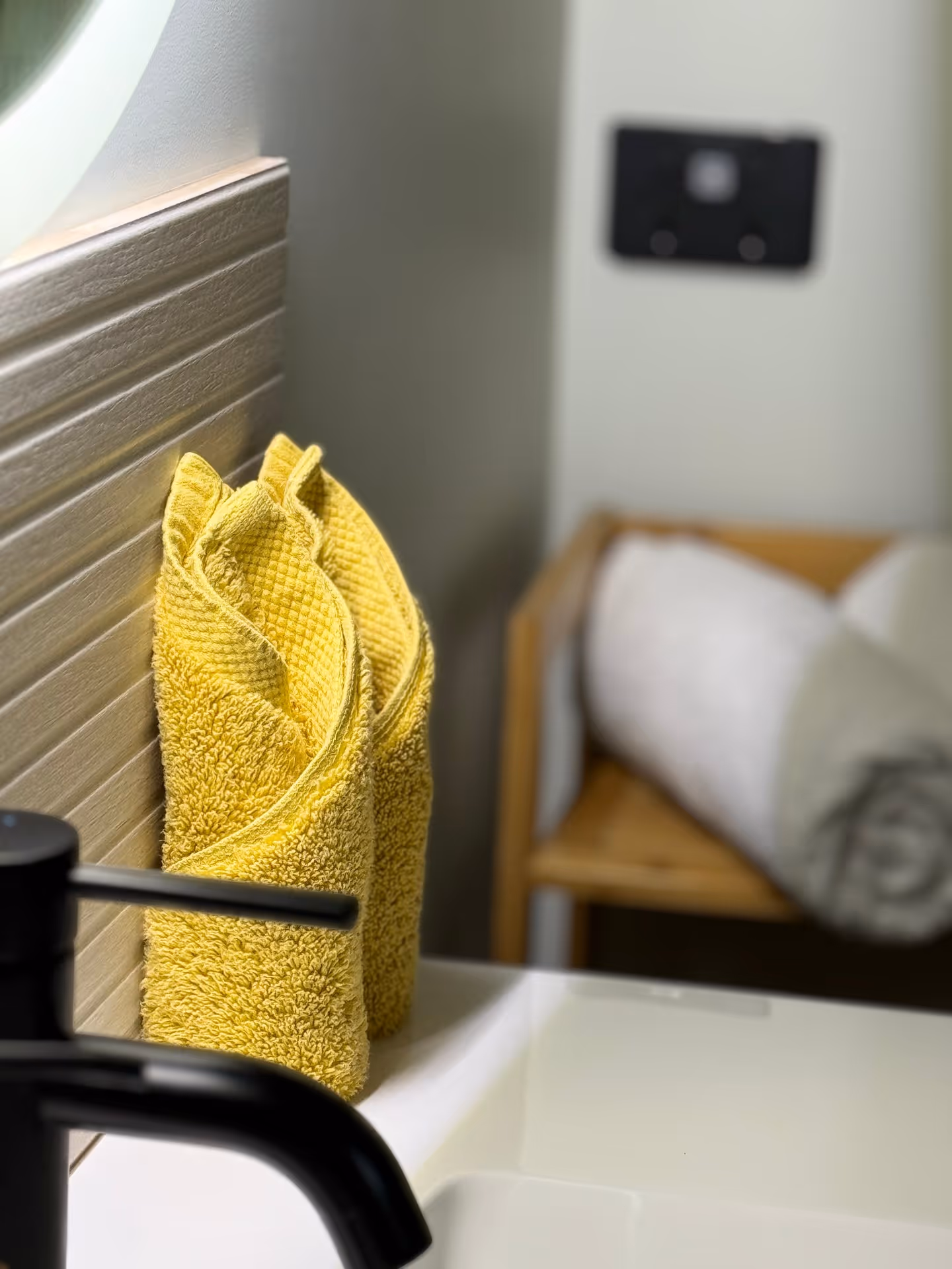Yellow towels are neatly folded beside a modern black faucet on a white countertop. A blurred white towel sits on a wooden rack, creating a serene bathroom vibe.