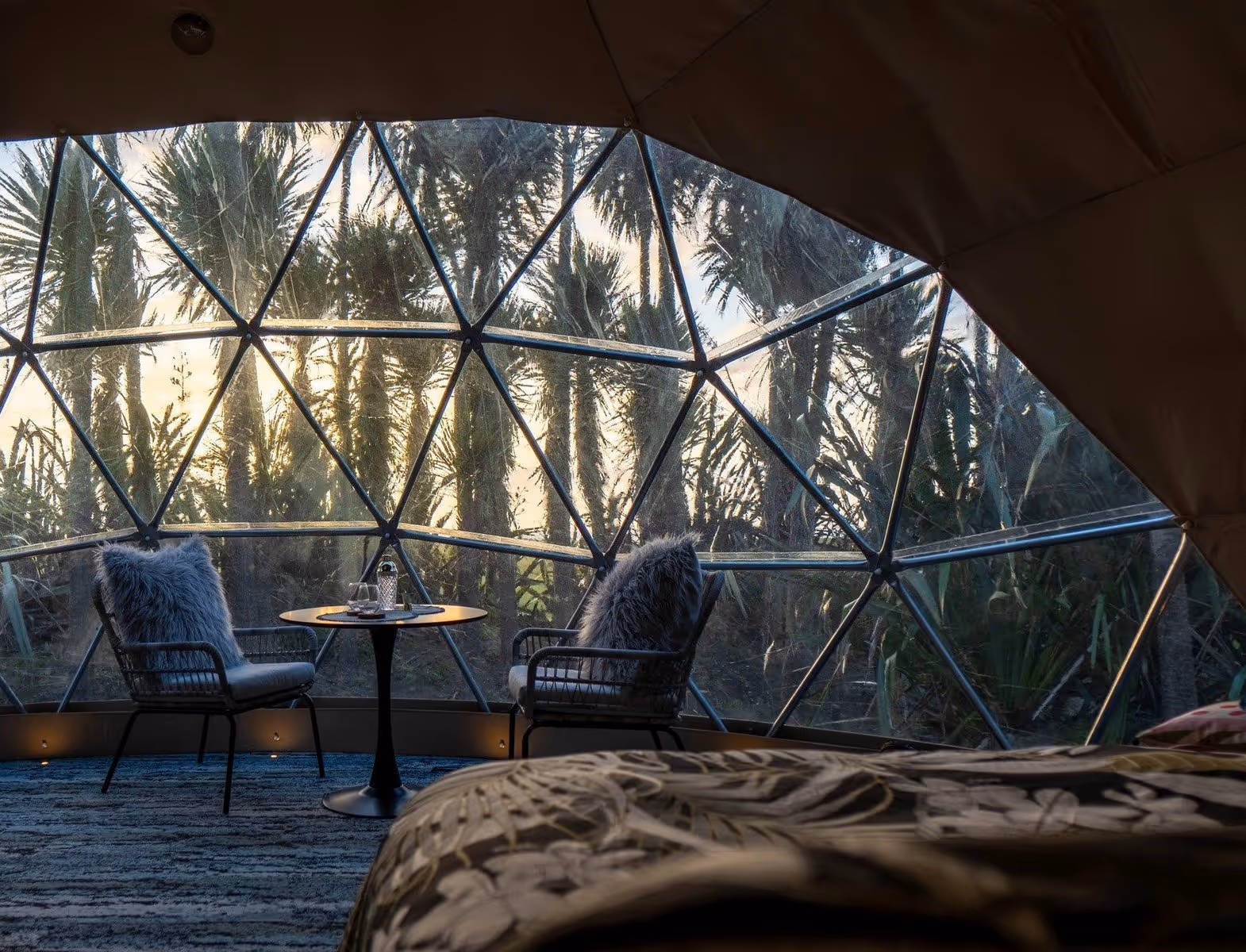 Interior of a geodesic dome with clear panels. Two chairs and a table face a forest at sunrise. Cozy and tranquil atmosphere, inviting relaxation.