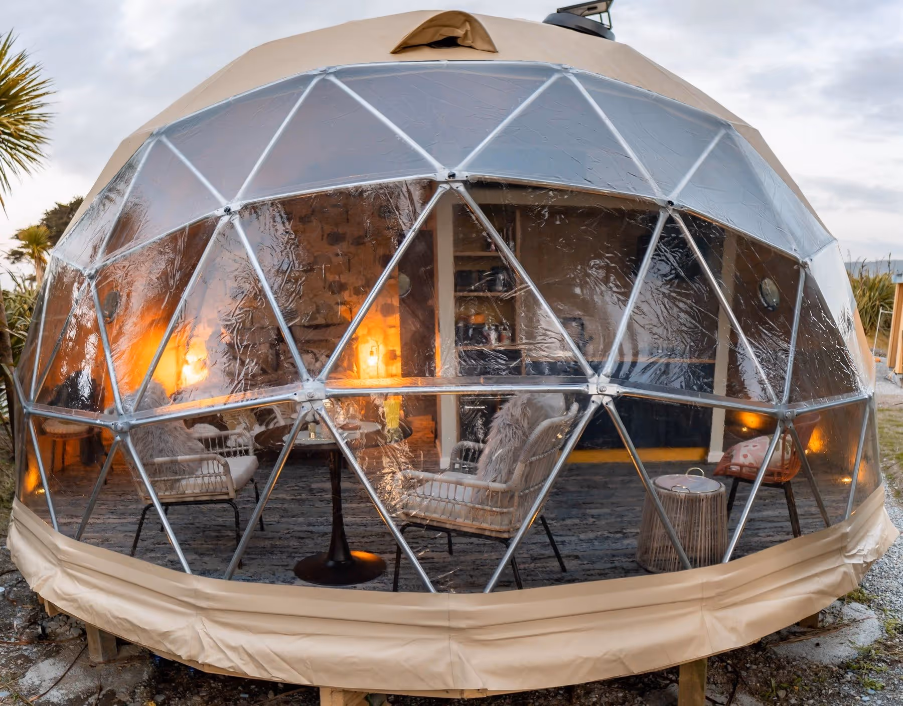 A cozy geodesic dome with transparent panels showing an inviting interior. Warm lights glow inside, surrounded by a serene, natural setting at dusk.