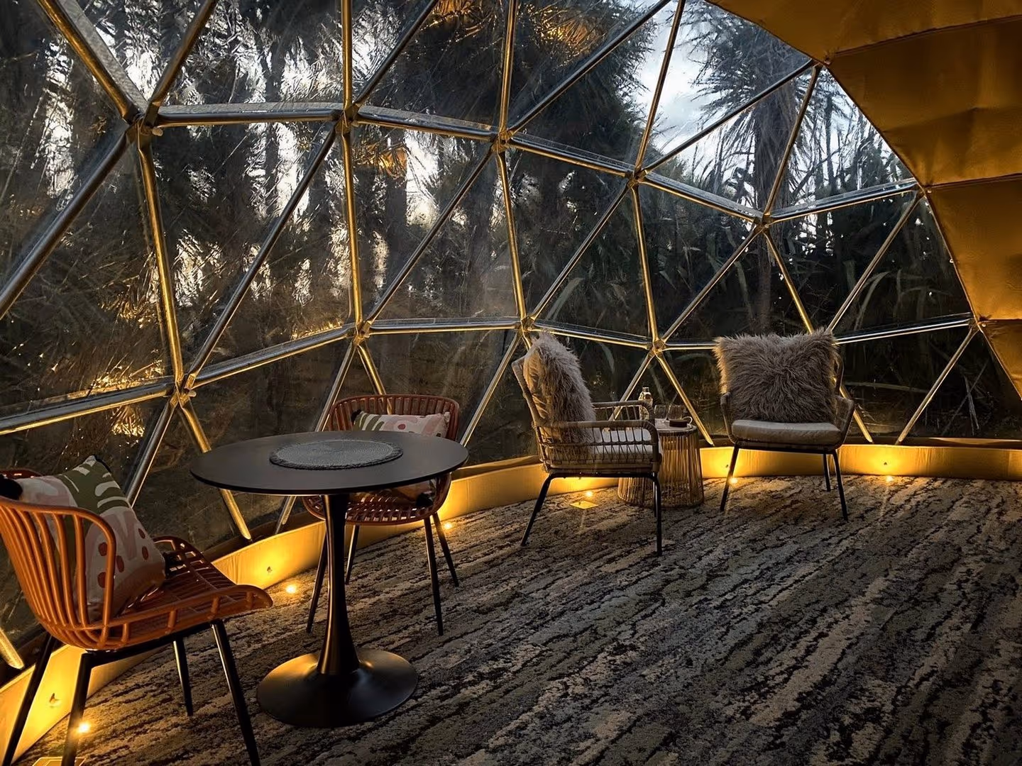 Cozy interior of a geodesic dome with warm lighting, featuring a table and chairs. Large windows reveal an exterior forest view at dusk.