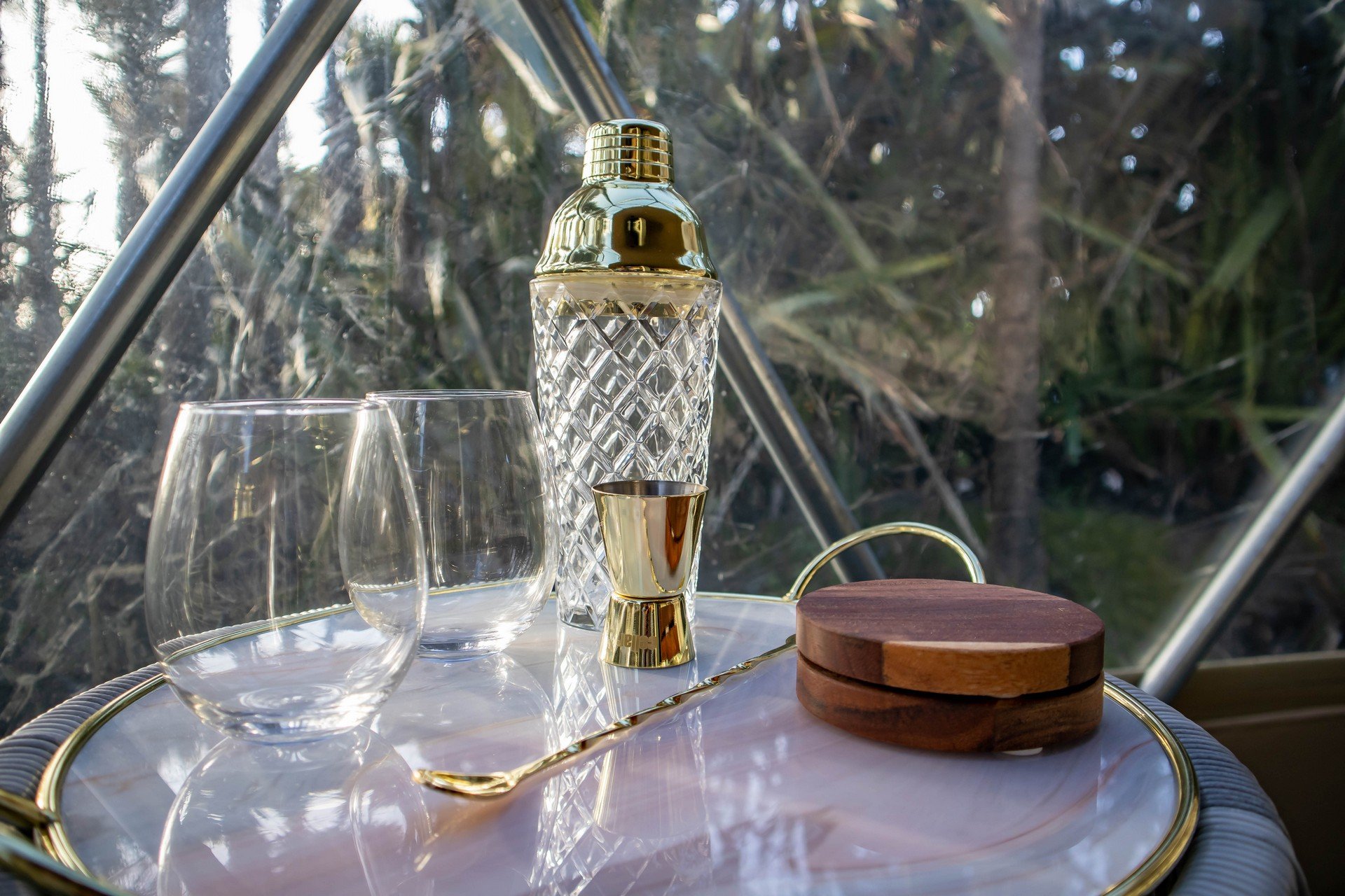 A serene table setting with two empty glasses, a gold-accented bottle, and a compact on a pink surface. Background shows trees through glass panels.