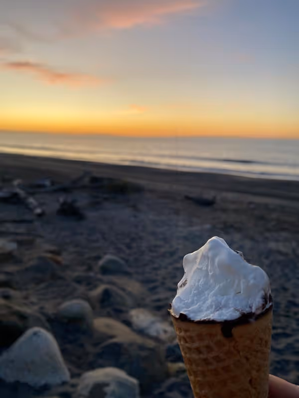Ice cream cone held on a rocky beach at sunset. The sky is painted with soft pink and orange hues, creating a serene and peaceful atmosphere.