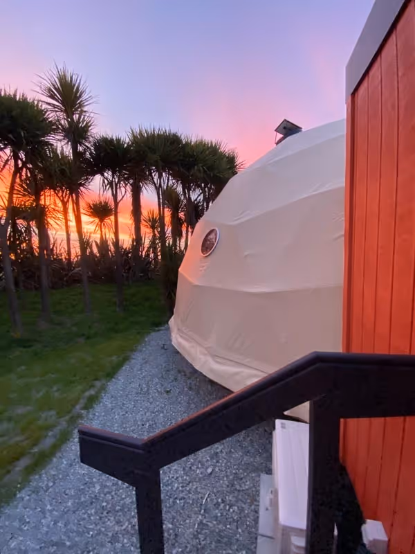 Sunset over trees with a warm glow in the sky, beside a modern dome-shaped tent on a gravel path, conveying a sense of tranquility and nature.