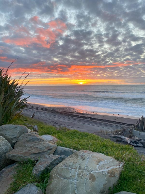 Sunset over a beach with a cloudy sky tinged with pink and orange hues. Waves gently lap the shore beside rocks and grass, creating a serene scene.