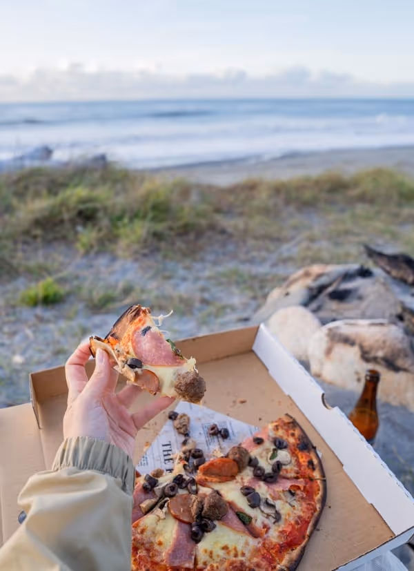 A hand holds a slice of pizza above an open pizza box on a beach. The ocean and sky are visible in the background, conveying a relaxed, coastal vibe.