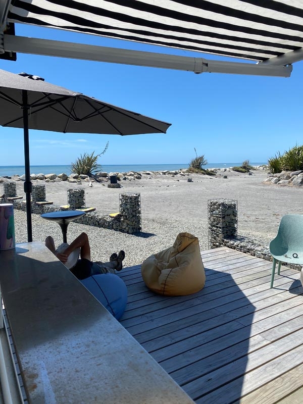 Outdoor patio with bean bags and a table under an umbrella on a wooden deck, overlooking a sandy beach and calm sea under a clear blue sky. Relaxing vibe.