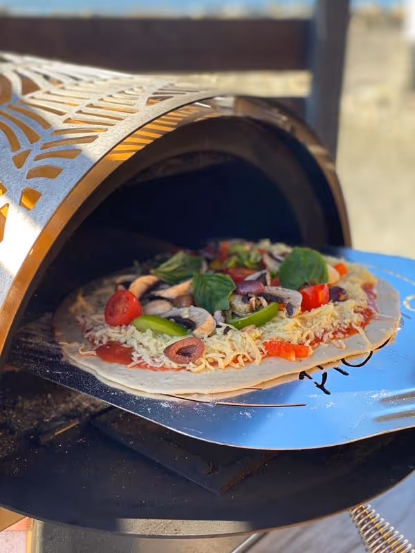 A homemade pizza with colorful toppings like bell peppers, mushrooms, and basil emerges from a hot, decorative oven onto a blue plate, suggesting warmth and freshness.