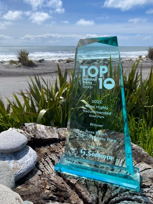A teal glass award inscribed with "Top 10" stands upright on a rocky surface, with a scenic beach and ocean in the background under a partly cloudy sky.