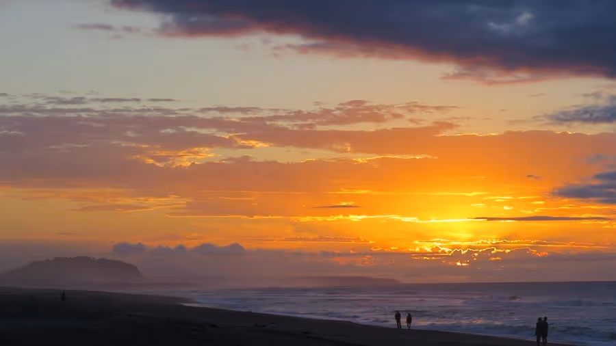 Sunset over a beach with vivid orange and yellow hues blending into clouds. Silhouettes of people and distant hills enhance the serene atmosphere.