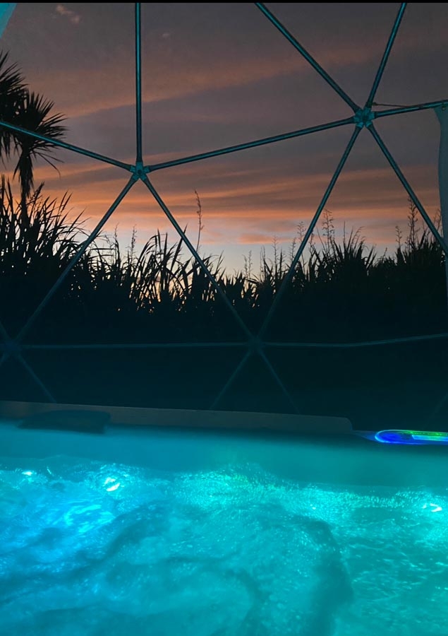 A tranquil pool with glowing blue water is in the foreground, enclosed by a geodesic dome. The sunset sky casts warm shades, with silhouettes of trees.