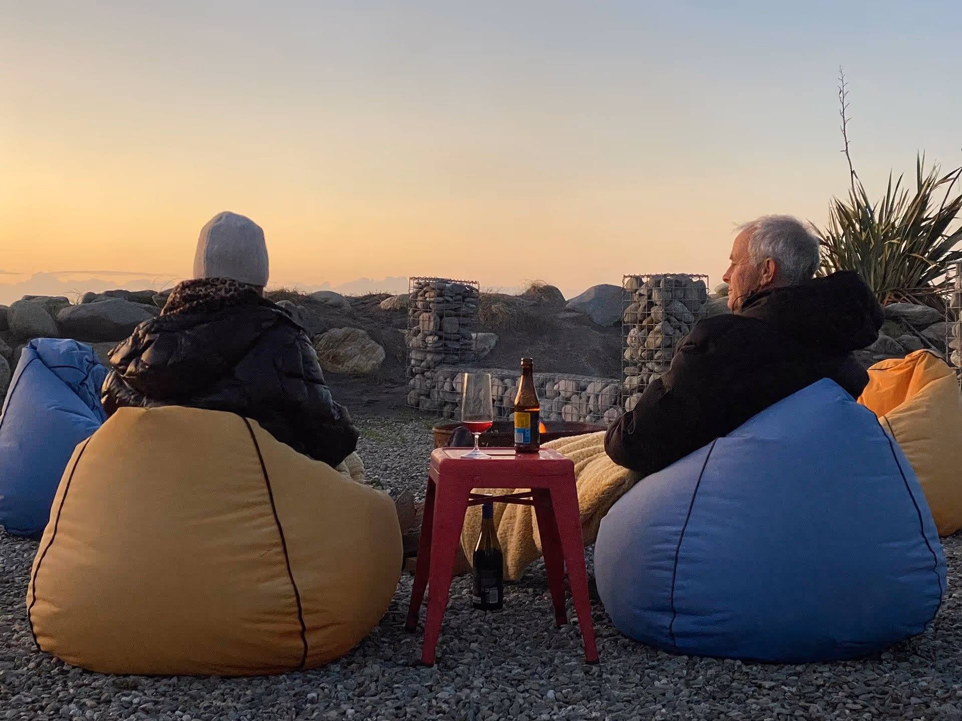 Two people sit on bean bags, facing a sunset. A small red table with drinks is between them. The scene conveys relaxation and tranquility.