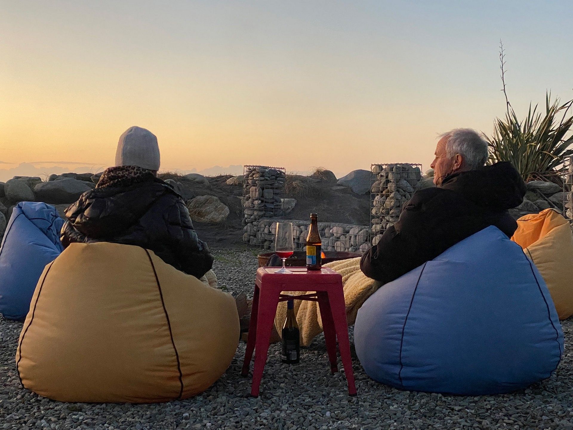 Two people sit on bean bags, facing a sunset. A small red table with drinks is between them. The scene conveys relaxation and tranquility.