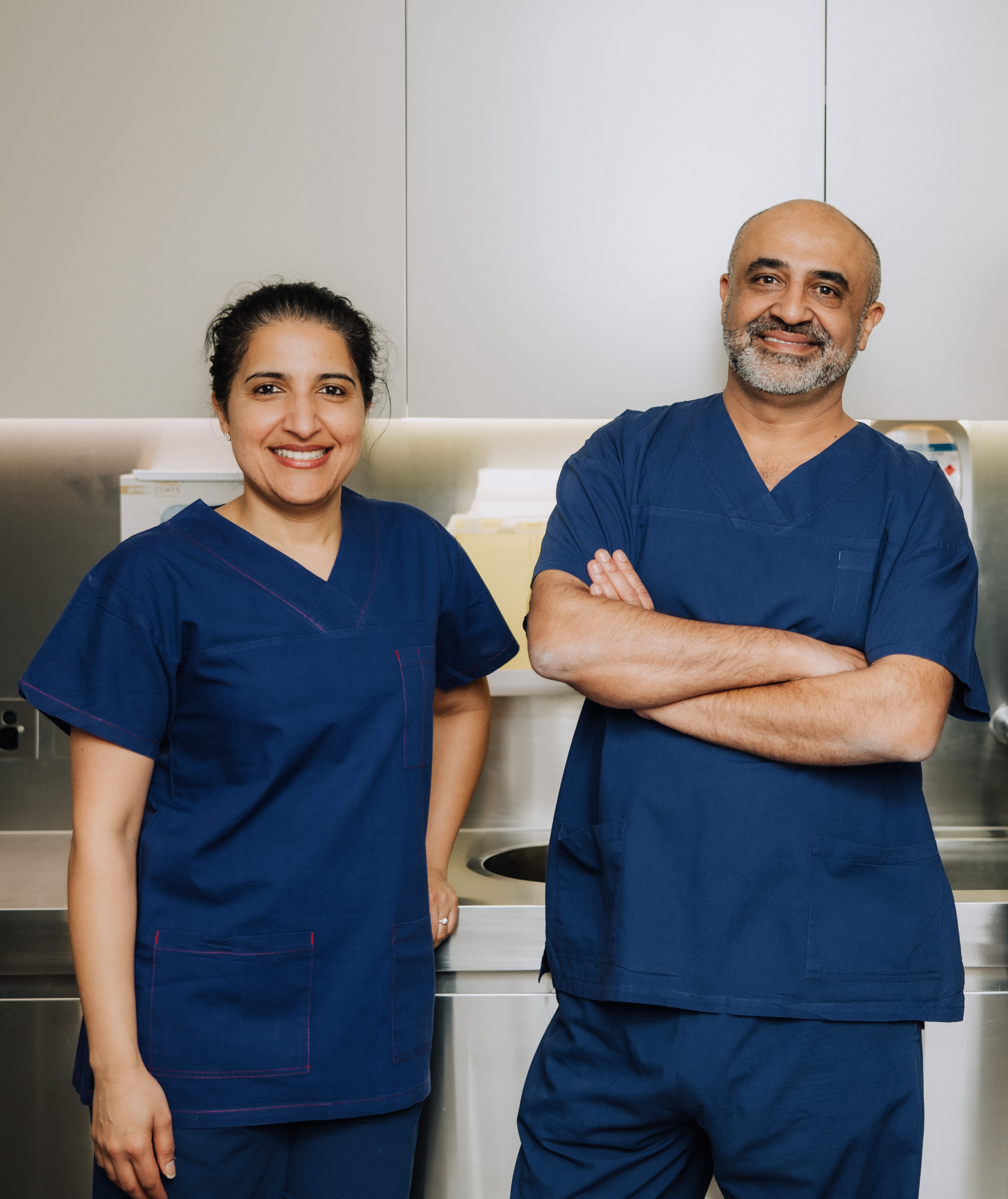 Two healthcare professionals in blue scrubs smiling in a clinical setting with stainless steel cabinets and sink.