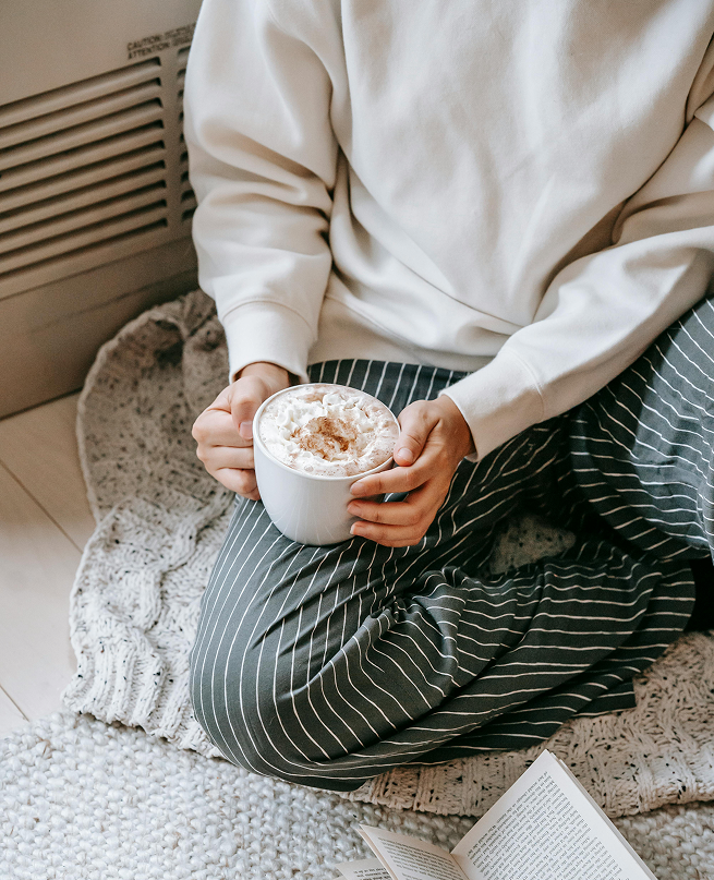 Person in striped pants and white sweatshirt sitting cross-legged on a knitted blanket, holding a mug topped with whipped cream and cinnamon.