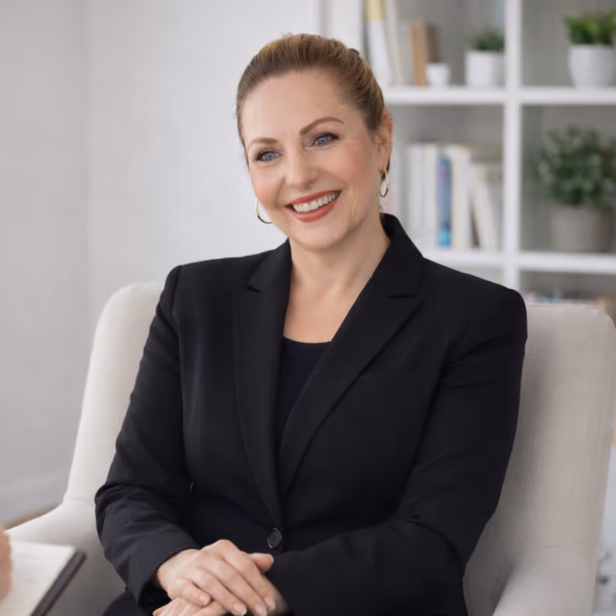 Lächelnde Frau in einem schwarzen Blazer sitzt auf einem hellen Stuhl in einem modernen Büro mit Bücherregalen hinter ihr.