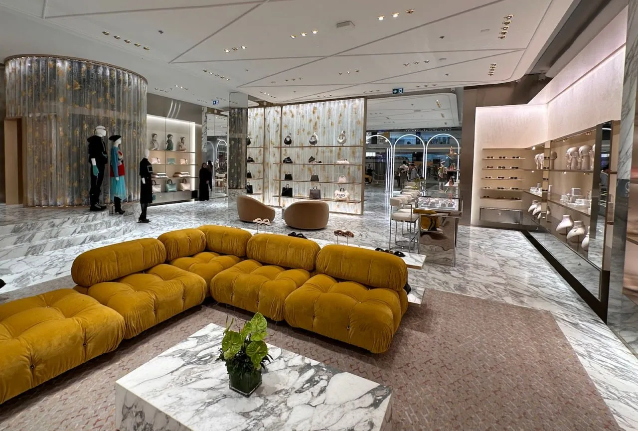 Luxurious retail store interior with mustard yellow sectional sofa, marble flooring, and displays of handbags, shoes, and mannequins.