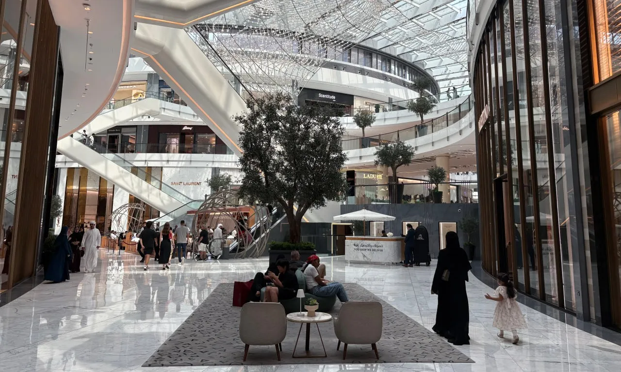Spacious modern shopping mall interior with marble floors, large tree, people walking, and multiple store levels connected by escalators.
