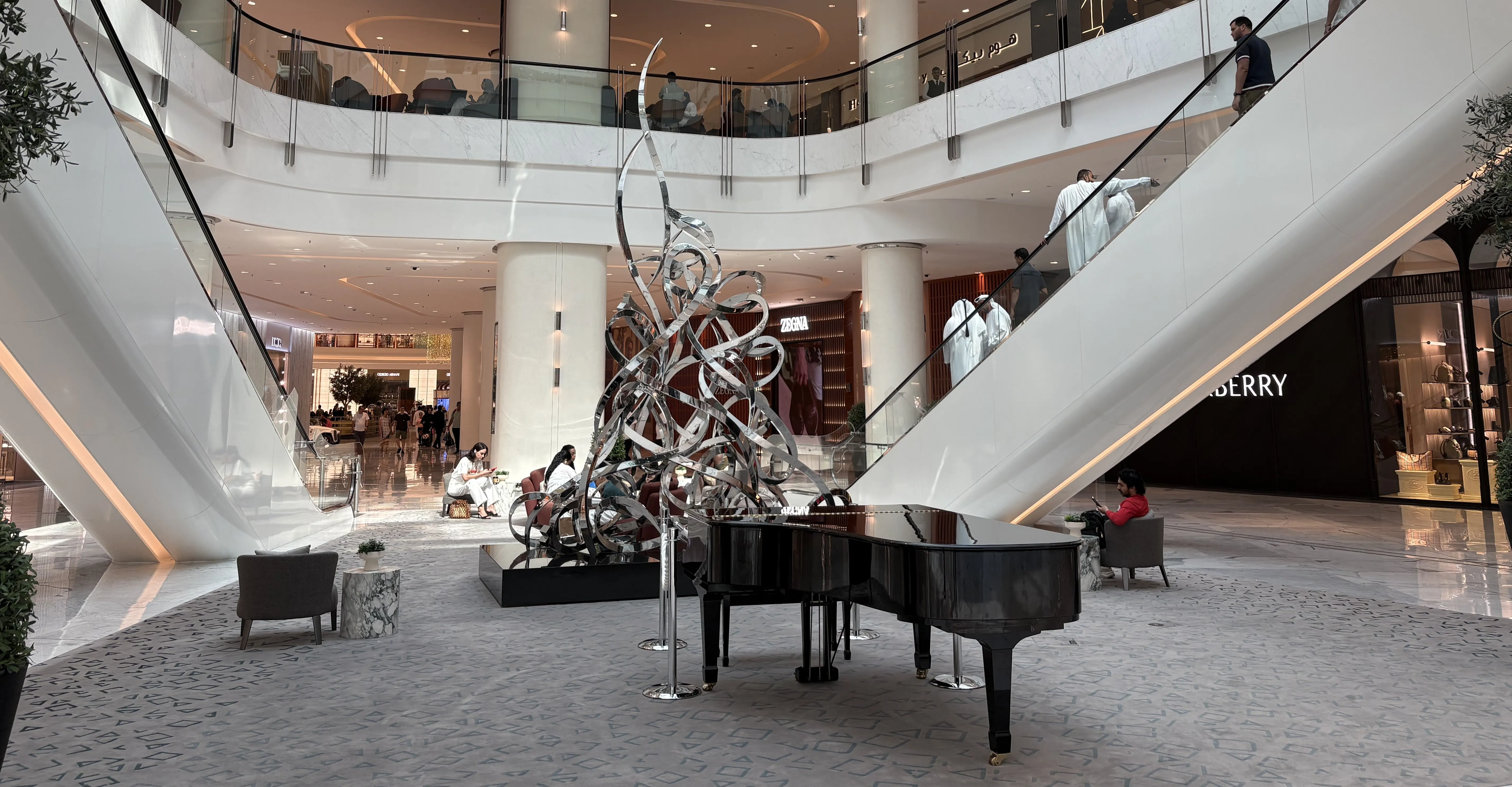 Modern shopping mall interior with a grand piano, abstract metal sculpture, escalators, and people sitting and walking.