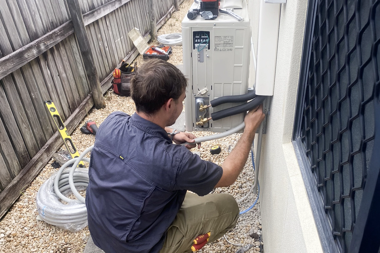A technician crouches beside an outdoor air conditioning unit, connecting insulated pipes and wiring on a gravel pathway next to a wooden fence.