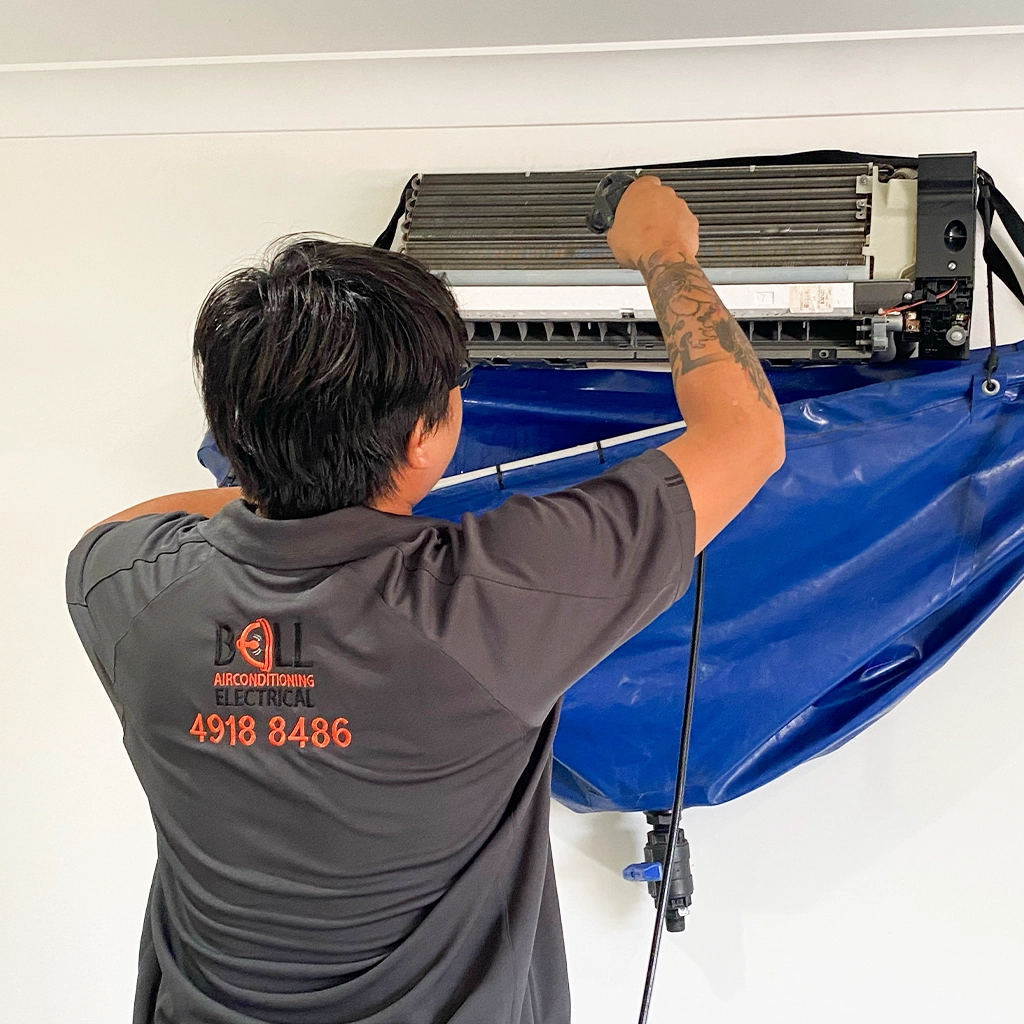 A technician cleans an indoor air conditioning unit using a spray tool, with a blue waterproof cover attached beneath to collect water and debris.