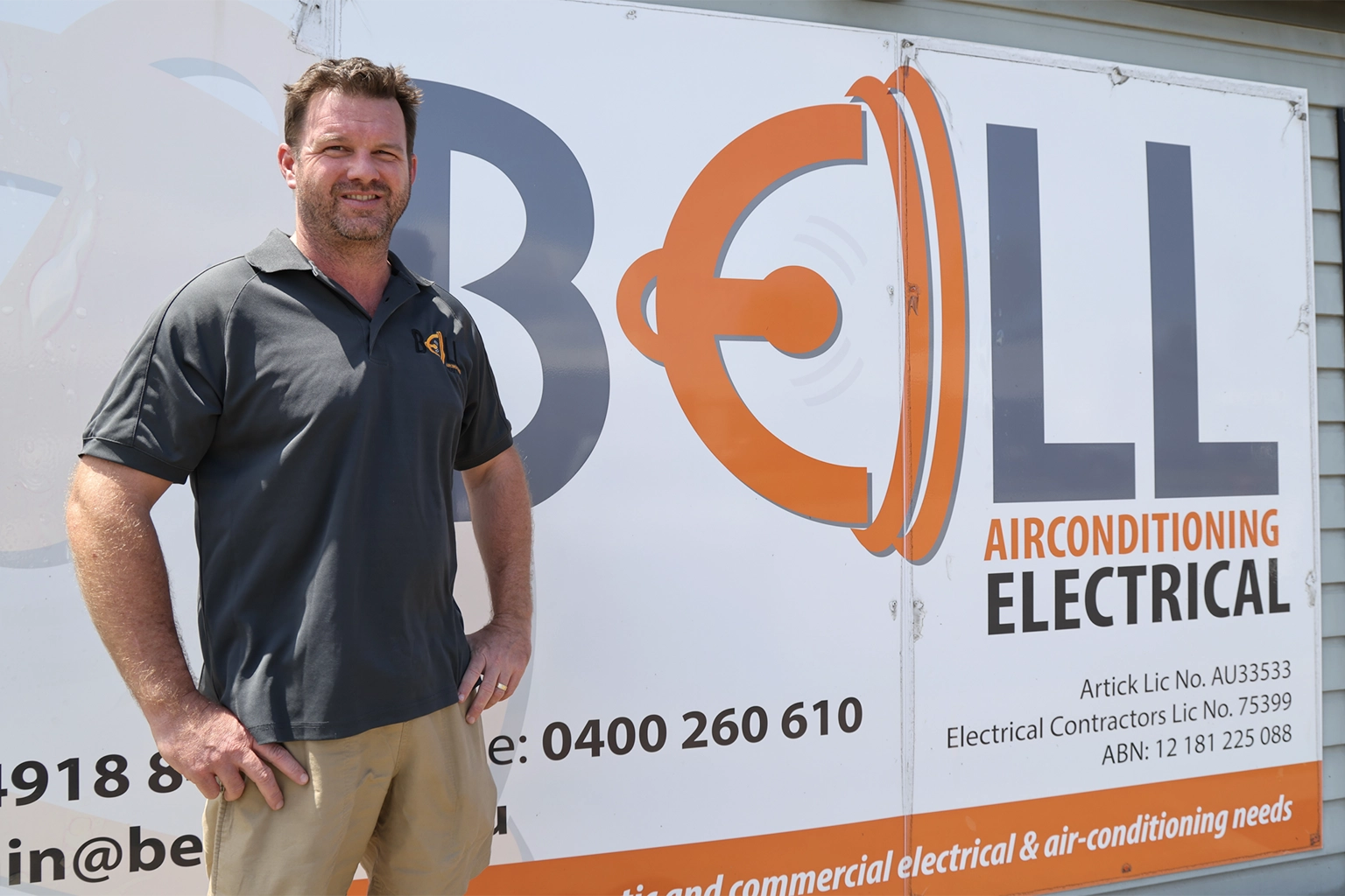 A smiling electrician from Bell Electrical installs or adjusts an outdoor wall light fixture on a brick house, using a screwdriver while standing in a neatly maintained backyard.