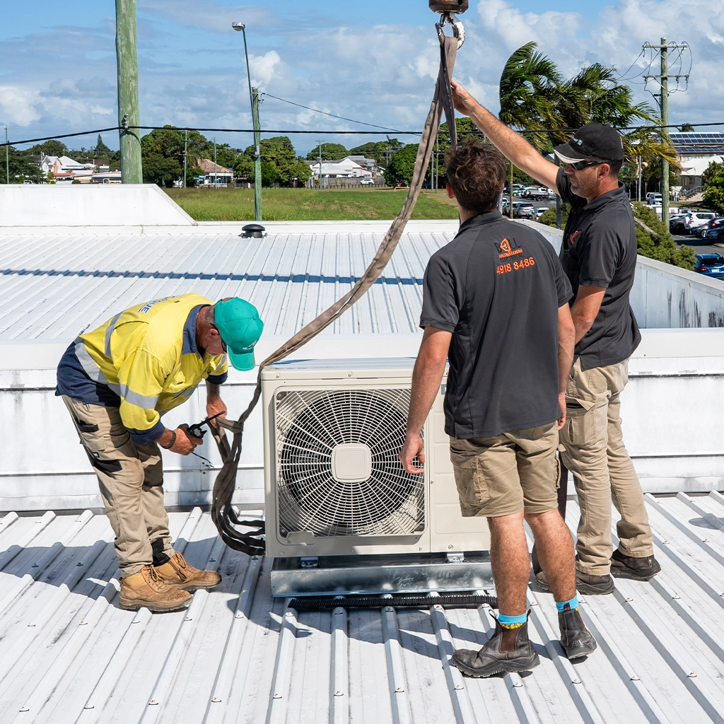 Two smiling technicians wearing black polo shirts and caps with the Bell Electrical logo stand on a rooftop beside an outdoor air conditioning unit on a sunny day.