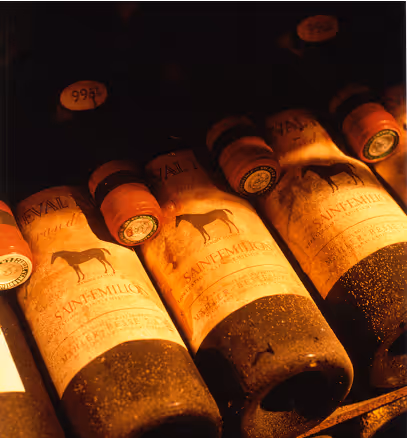 Bouteilles de vin Saint-Emilion poussiéreuses alignées sur une étagère en bois, éclairées par une lumière chaude.