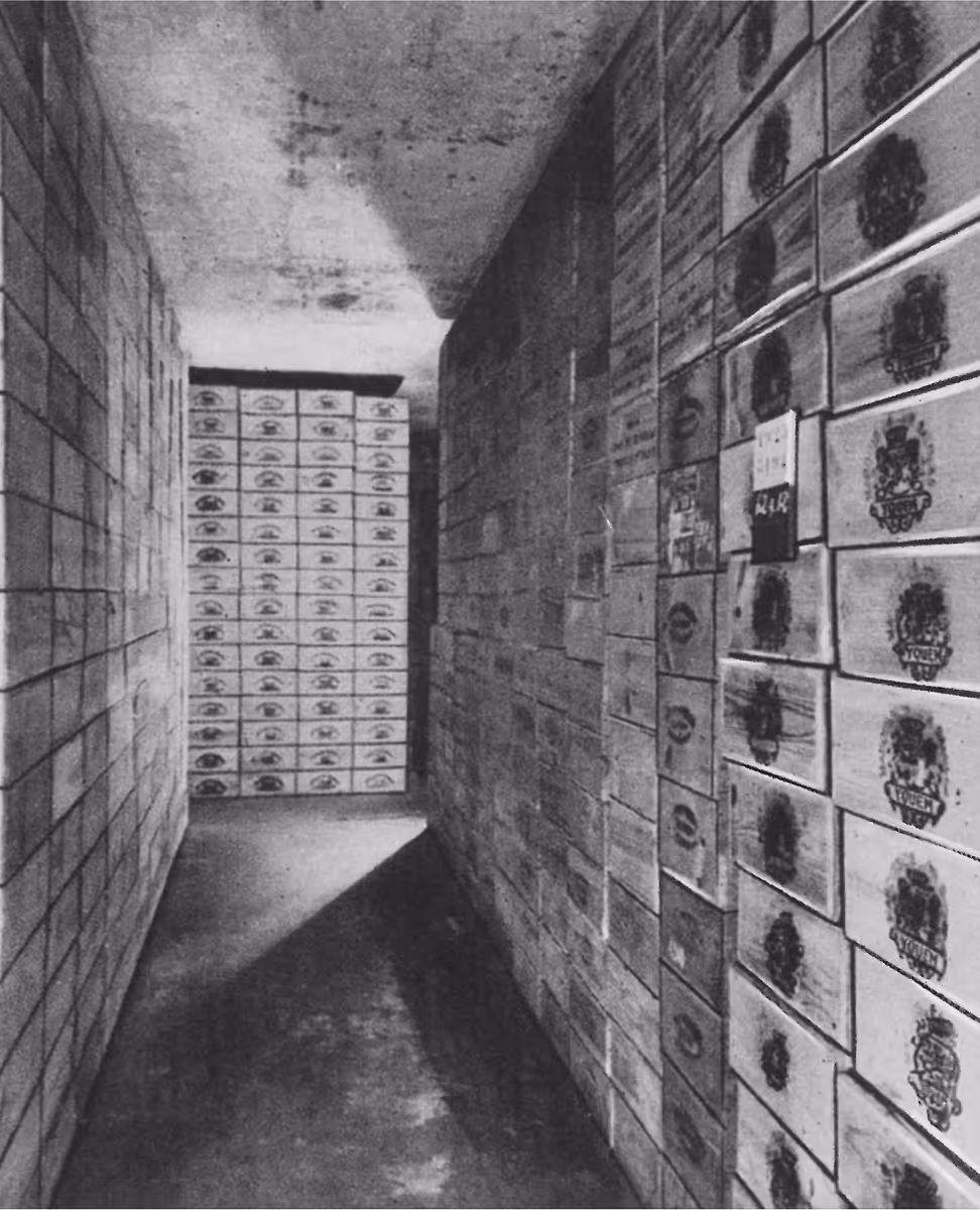 Couloir sombre avec des murs couverts de caisses en bois empilées, certaines portant un blason.