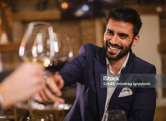 Homme souriant en veste bleue trinquant avec un verre de vin rouge dans un cadre chaleureux.