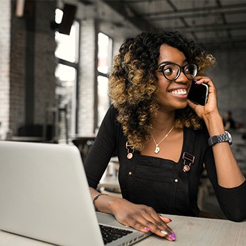 Jeune femme souriante portant des lunettes, parlant au téléphone avec un ordinateur portable devant elle dans un bureau moderne.