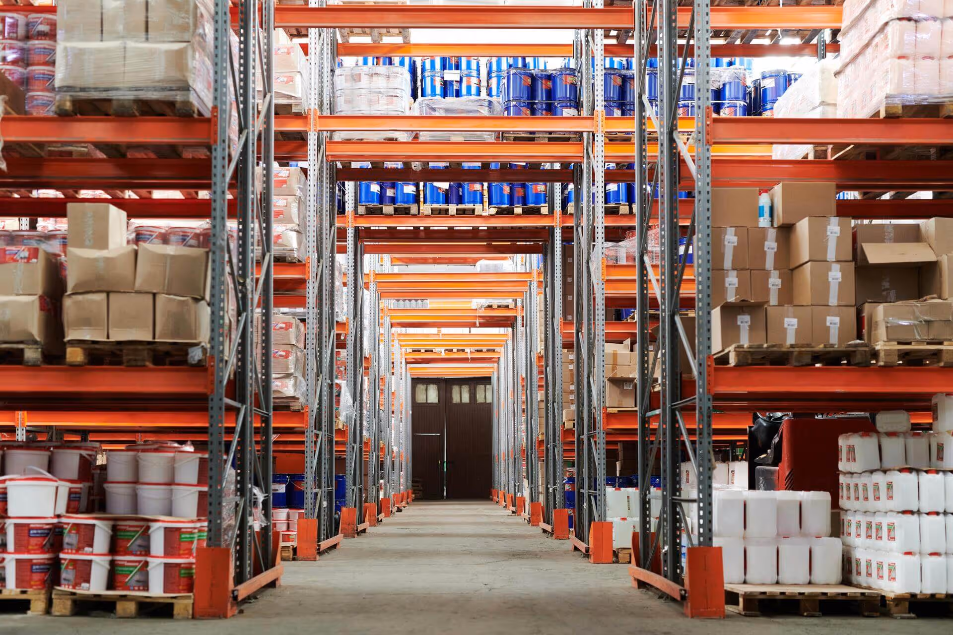Aisle in a warehouse with orange metal shelving stocked with various boxes, white plastic containers, and blue barrels.