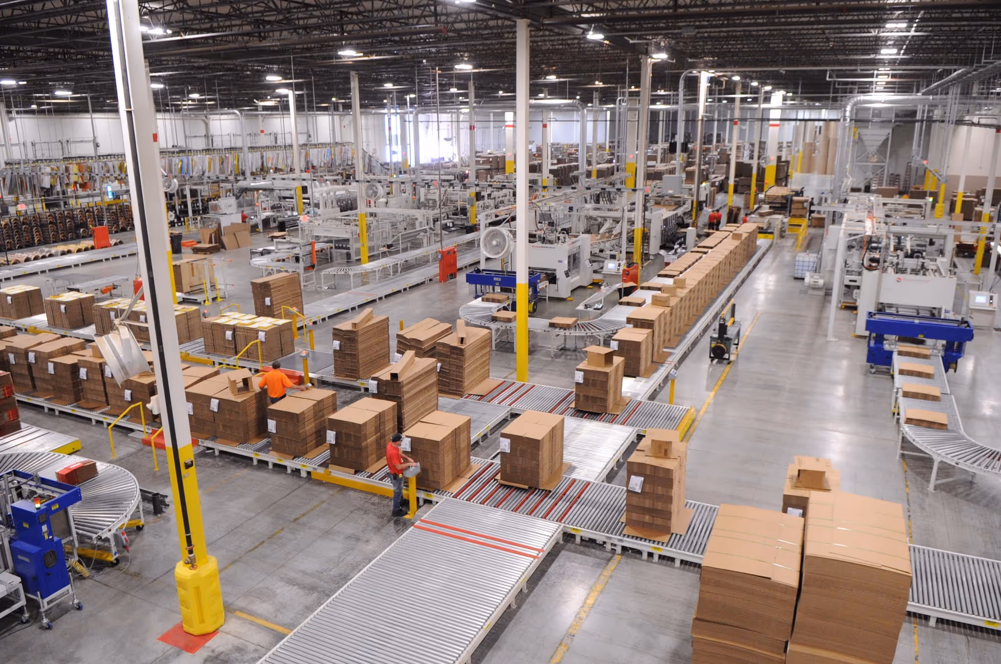 Large warehouse with conveyor belts transporting stacked cardboard boxes and workers managing packaging.
