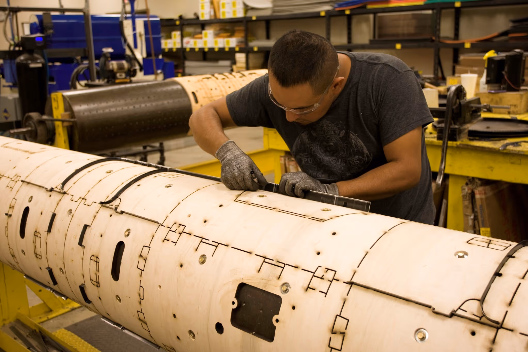 A man wearing gloves and safety glasses works on assembling a large cylindrical wooden structure with detailed markings in a workshop.