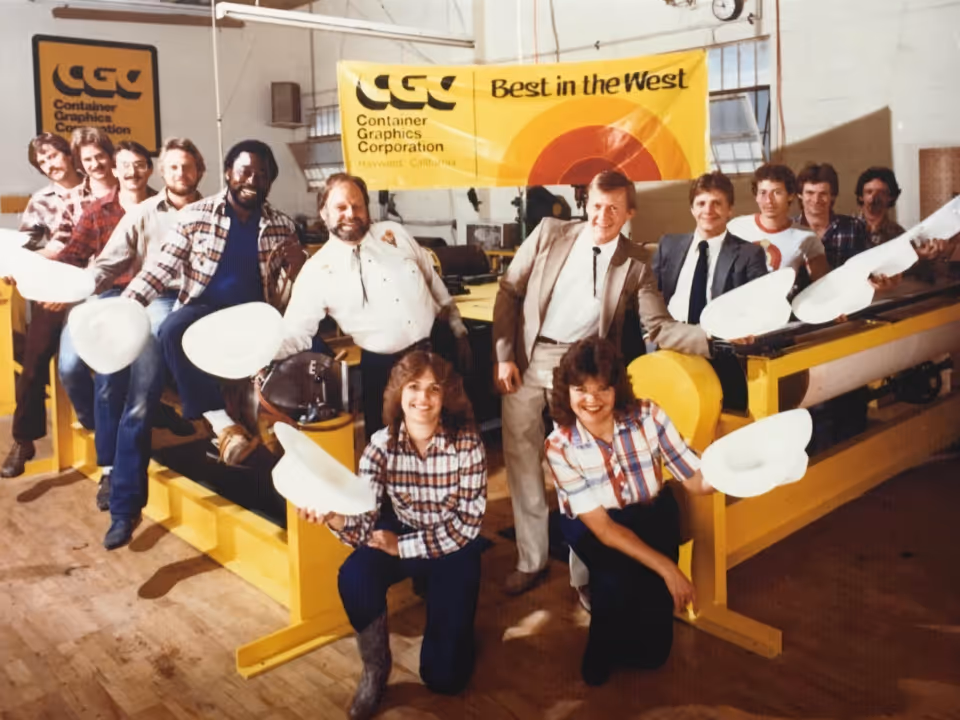 Group of people indoors holding white hats, posing in front of yellow equipment and banners reading CGC Container Graphics Corporation and Best in the West.
